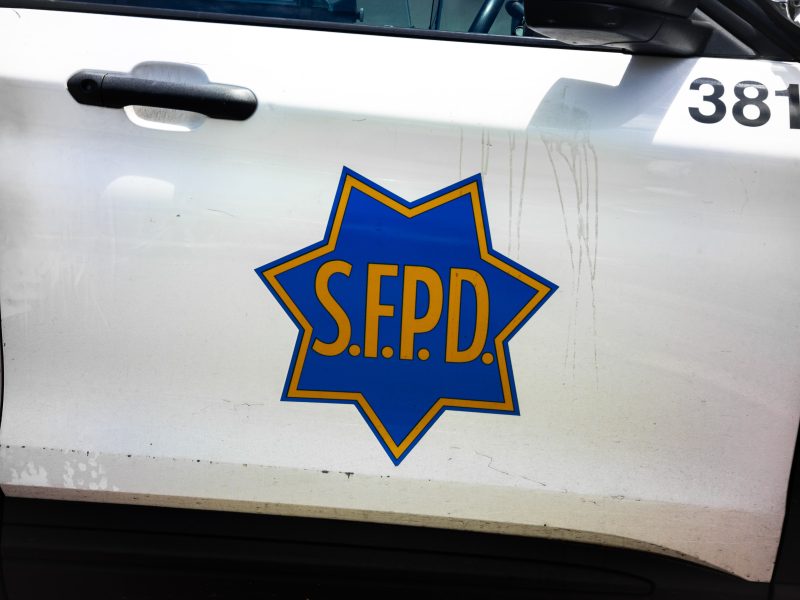 Side view of a white police vehicle with a blue and yellow S.F.P.D. badge logo on the door and the number 381 above.