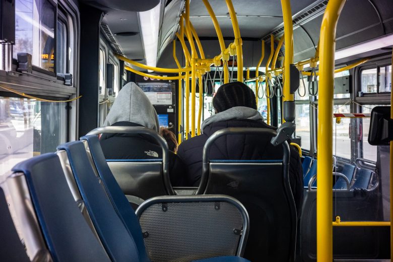 Two people wearing hooded jackets sit near the back of a city bus with yellow handrails and blue seats, facing forward.