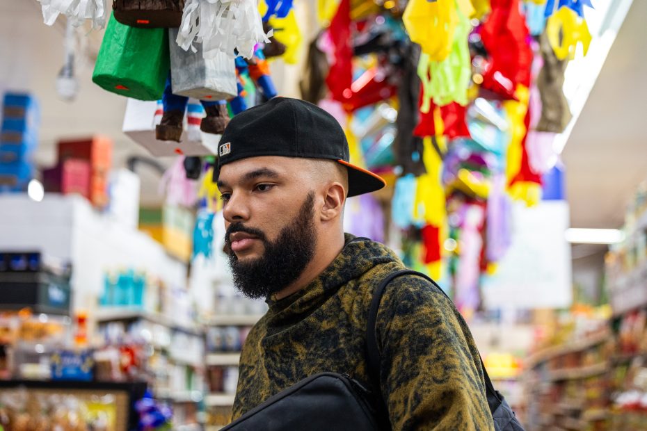 A man wearing a black cap and green hoodie stands in a store aisle with colorful hanging decorations overhead.