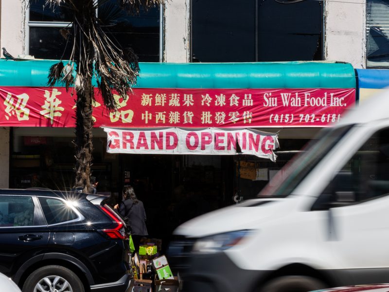 A storefront with a green and red awning displays "Sit Wah Food Inc." and a large "Grand Opening" banner; cars pass by on the street in front.