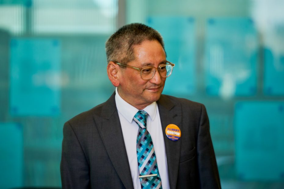 A man in a suit and glasses stands indoors, wearing a turquoise patterned tie and a button that reads "Albert Chow."