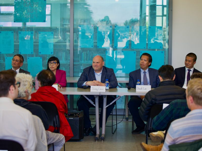 Five panelists sit at a table facing an audience during a discussion event, with nameplates and water bottles in front of them.
