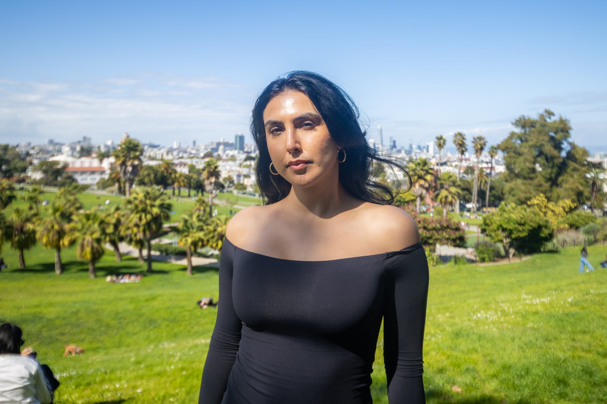 A person in a black off-shoulder dress stands in a sunny park with palm trees and city buildings visible in the background.