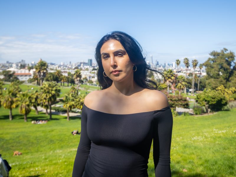 A person in a black off-shoulder dress stands in a sunny park with palm trees and city buildings visible in the background.