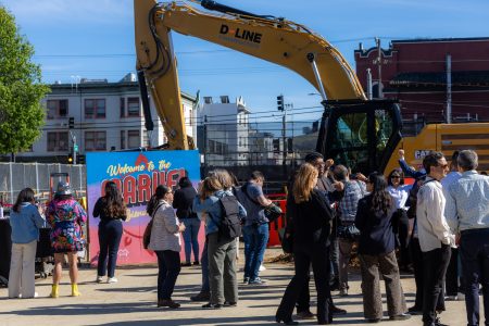 A group of people gather near a construction site with a large excavator and a sign that reads “Welcome to the Barrio.” Buildings and a clear sky are visible in the background.
