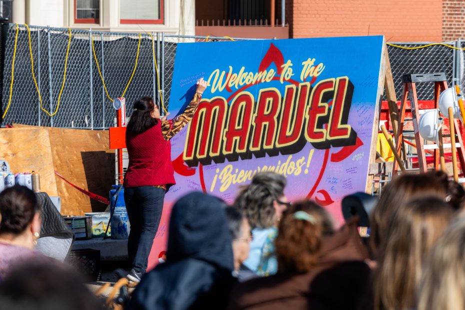 A woman writes on a large sign that reads "Welcome to the Marvel ¡Bienvenidos!" while a seated crowd watches at an outdoor event.