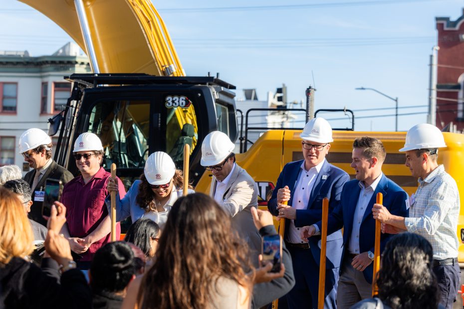 A group of people in hard hats stand in front of construction equipment at an outdoor event, holding ceremonial shovels while a crowd looks on.