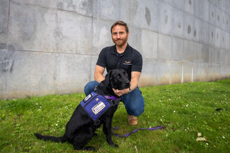 A man kneels on grass beside a black dog wearing a service vest, in front of a gray stone wall.