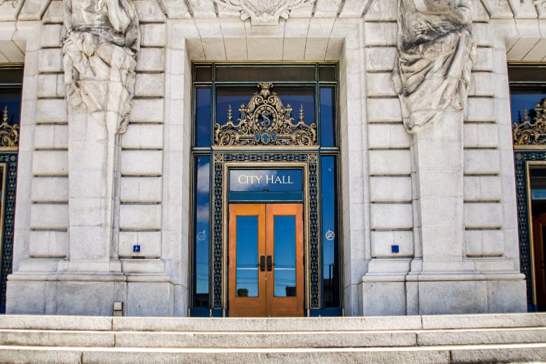 Stone steps lead to ornate double doors with a sign above reading "City Hall" on a grand, detailed building facade.