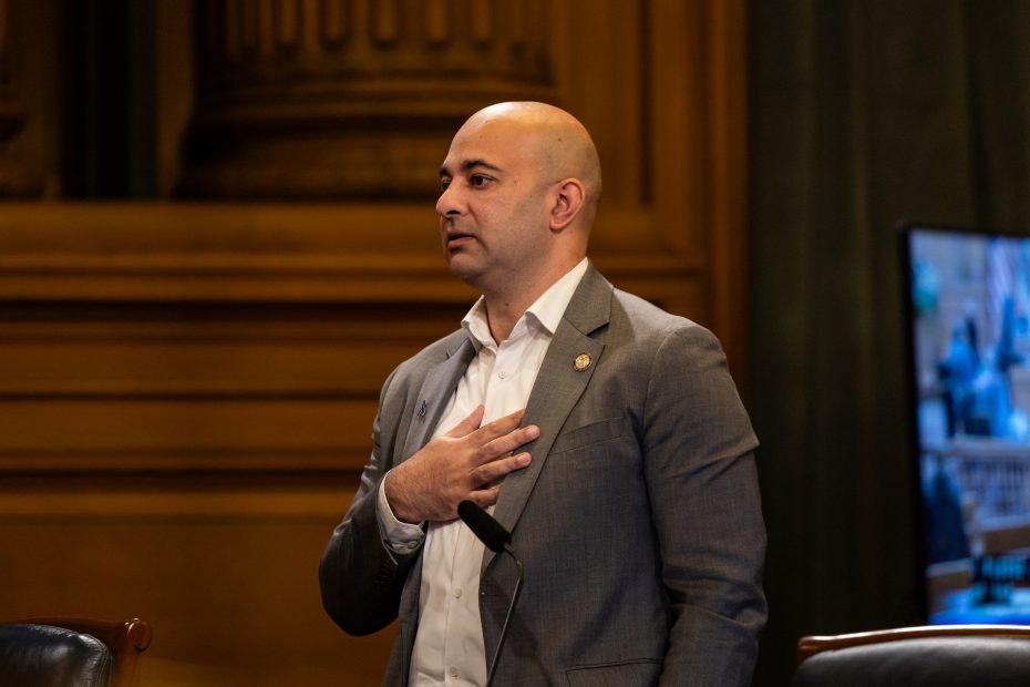 A man in a suit stands indoors with his hand on his chest, speaking or presenting in a formal setting with ornate wooden details in the background.