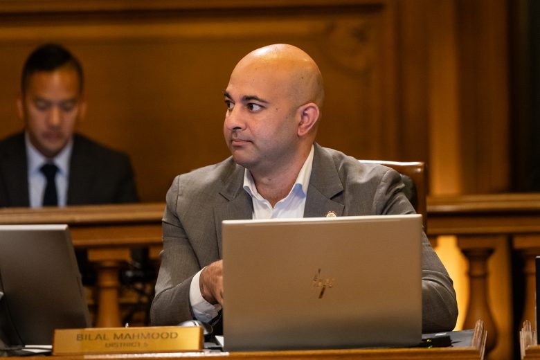 A man in a suit sits at a desk with a laptop in a formal meeting room. Another person sits in the background. A nameplate reads "Bilal Mahmood.