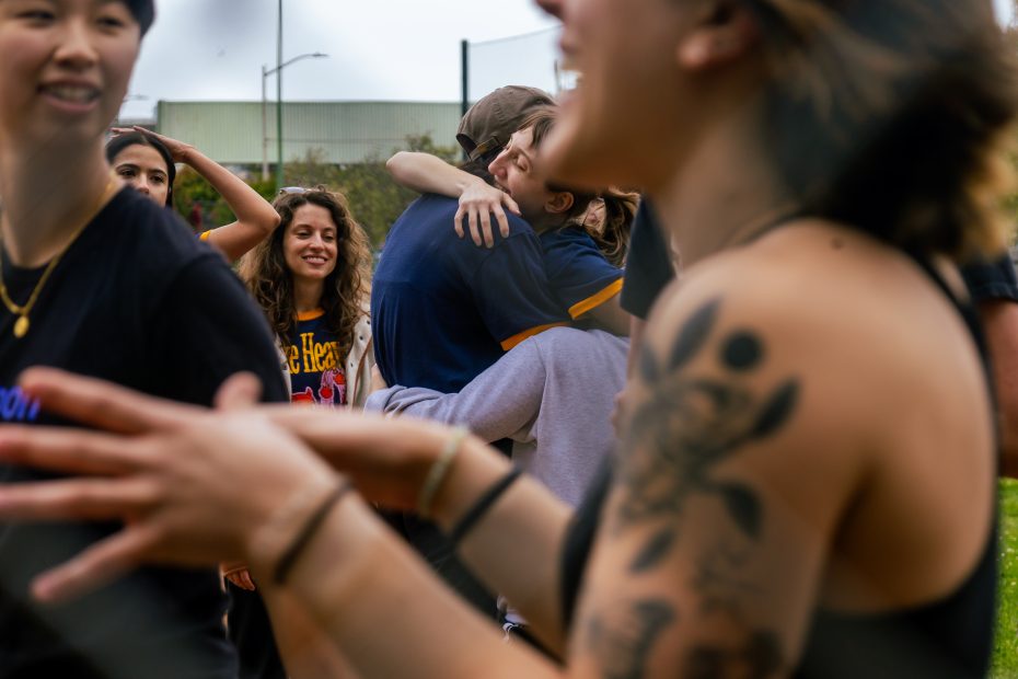 A group of people gathers outdoors; two people hug in the background while others smile and talk in the foreground.