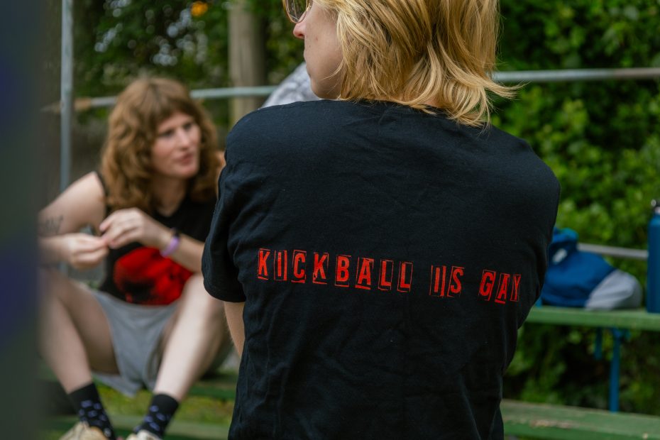 A person with blonde hair wears a black shirt with red text reading "KICKBALL IS GAY," sitting outside near another person on green bleachers.
