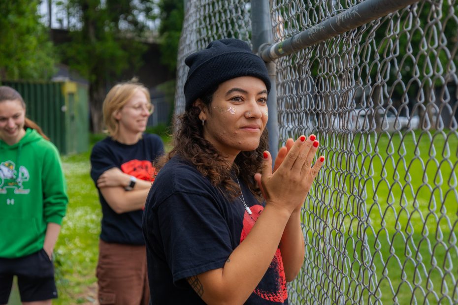 A woman in a black beanie stands by a chain-link fence with her hands clasped, while two people stand in the background on a grassy field.