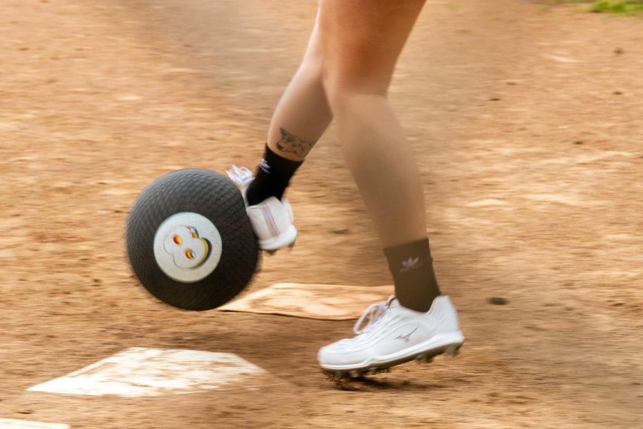 A person wearing white athletic shoes is kicking a round object with a smiling face printed on it on a dirt surface.