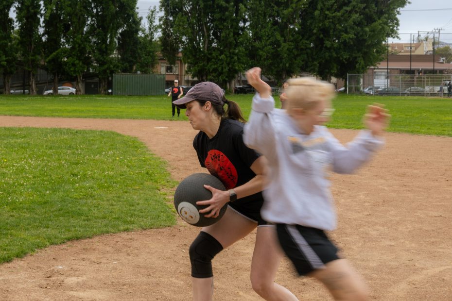 Two people are playing kickball on a dirt field; one person holds the ball while another runs past them in a blurred motion. Trees and buildings are visible in the background.