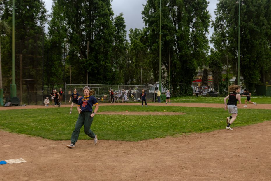 People playing a baseball game on a field, with one person running toward a base and others positioned around the diamond; trees and spectators are visible in the background.