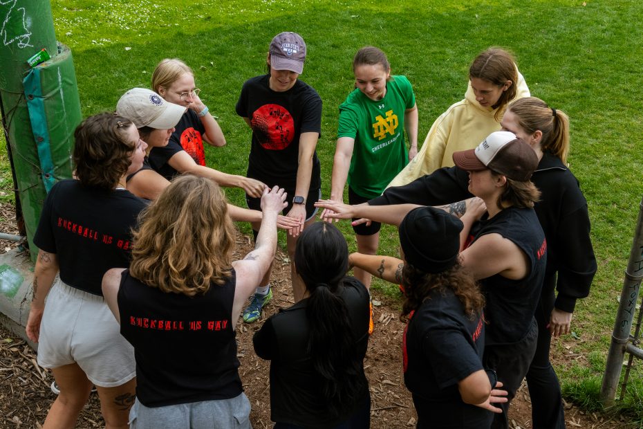 A group of people standing in a circle outdoors, each with one hand extended into the center, preparing for a team activity.