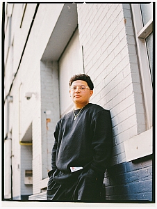A person wearing glasses and a black outfit stands against a light-colored brick wall, looking at the camera with hands in pockets.