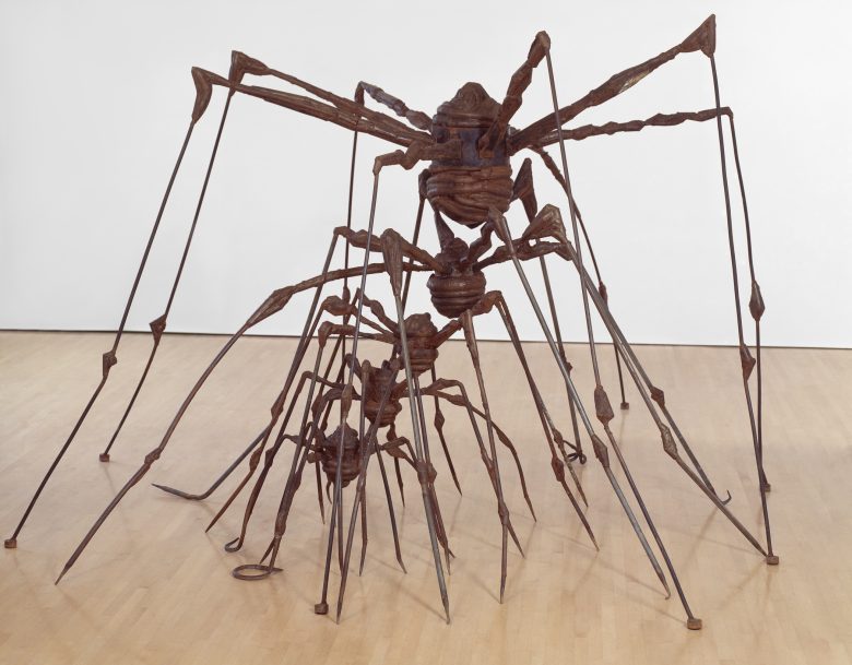 Large rust-colored metal sculpture of three spiders, each with elongated legs, displayed on a light wooden floor against a plain white wall.