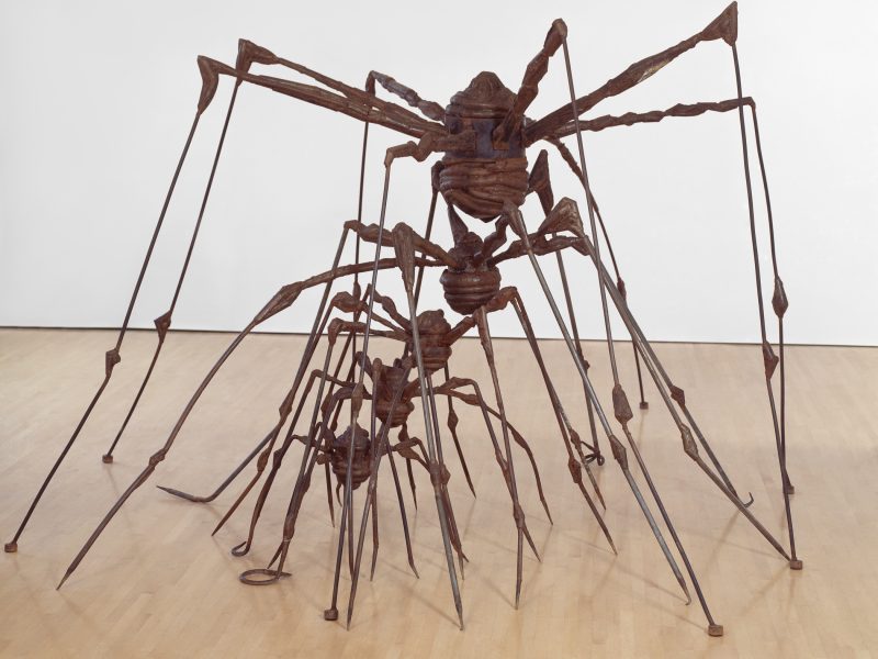 Large rust-colored metal sculpture of three spiders, each with elongated legs, displayed on a light wooden floor against a plain white wall.