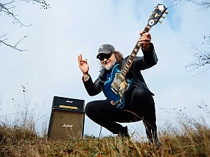 A person wearing sunglasses and a cap plays an electric guitar outdoors next to a Marshall amplifier, surrounded by dry grass and a clear sky.