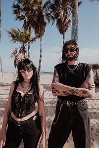Two people wearing black clothing and sunglasses stand side by side on a sandy beach with palm trees in the background.