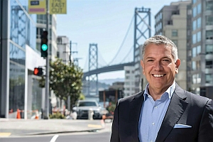 Smiling man in a suit stands on a city street with the Bay Bridge and modern buildings in the background.