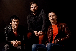 Three men in jackets sit and pose against a dark background, looking directly at the camera.