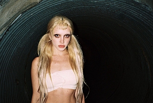 A woman with long blonde hair and dark eye makeup stands in front of a dark, ribbed tunnel, wearing a light bandeau top.