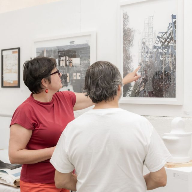 Two people stand in front of a framed architectural photograph in a gallery; one points at the image while discussing it with the other.