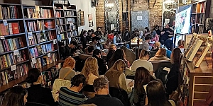 A group of people sits closely together in a bookstore, facing a projected screen, with bookshelves lining the walls.
