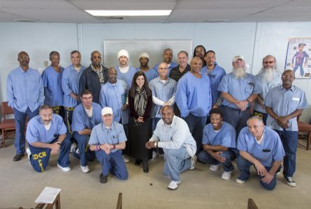 A group of men, mostly wearing blue uniforms, and one woman stand and kneel together in a classroom with chairs and a whiteboard.
