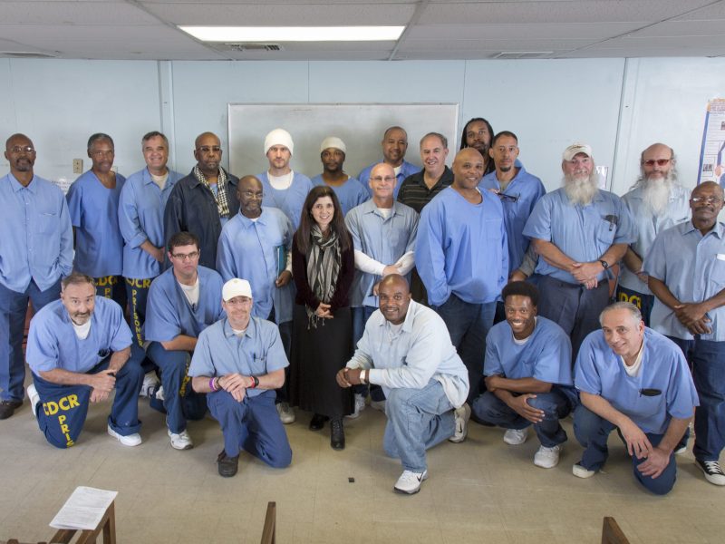 A group of men, mostly wearing blue uniforms, and one woman stand and kneel together in a classroom with chairs and a whiteboard.