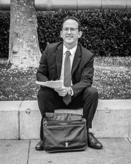 A man in a suit sits on a stone ledge outdoors, holding papers and smiling, with a briefcase at his feet and trees and bushes in the background.