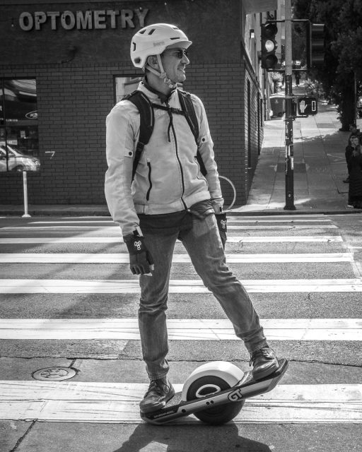 A man wearing a helmet and backpack stands on a one-wheeled electric skateboard at a crosswalk near an optometry clinic.