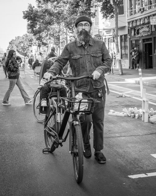 A man with a beard and glasses walks a bicycle down a city street during what appears to be a public event; other people and buildings line the street.