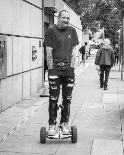 A man with tattoos and ripped jeans rides a two-wheeled self-balancing scooter on a city sidewalk; other pedestrians walk nearby.