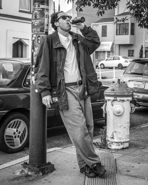 A man in sunglasses leans against a pole on a city sidewalk, drinking from a bottle near parked cars and a fire hydrant.