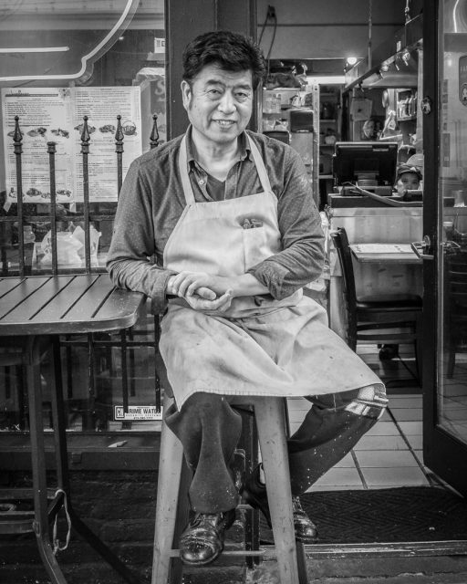 A man wearing an apron sits on a stool outside a restaurant, with menus visible in the background. The photo is in black and white.