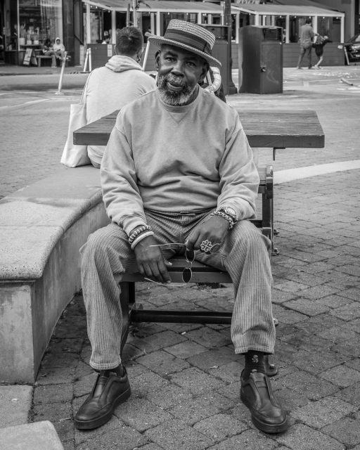 A man wearing a hat, long-sleeve shirt, and corduroy pants sits on a bench in an urban outdoor setting, looking at the camera. The image is in black and white.