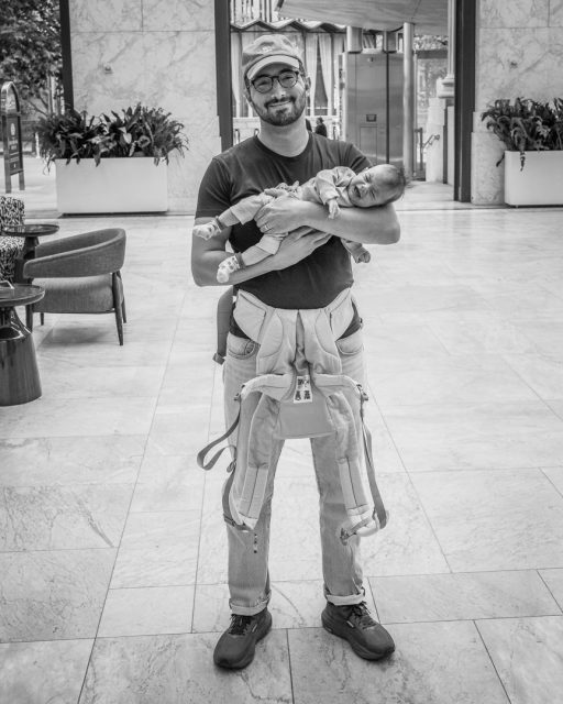A man wearing glasses and a cap stands indoors, holding a baby horizontally in his arms; an empty baby carrier hangs from his shoulders.