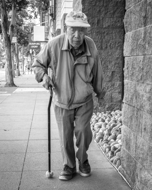 An elderly man wearing a jacket and cap walks with a cane on a city sidewalk next to a stone wall lined with rocks.