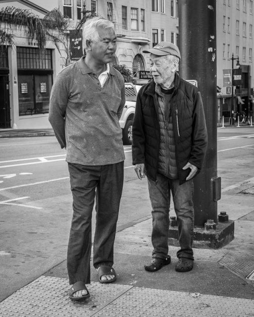 Two older men stand and talk on a city sidewalk; one has his hands behind his back, and both are dressed casually. Buildings and a street are visible in the background.