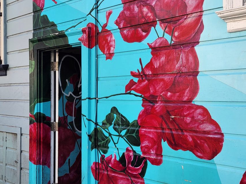A mural of large red flowers with green leaves painted on a blue wooden exterior wall, partially covering a doorway.