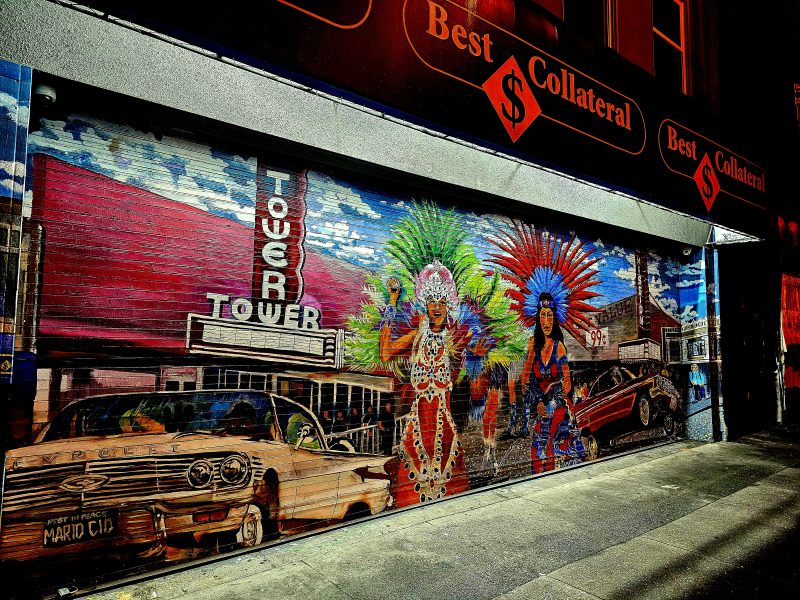 A colorful mural on a storefront depicts a vintage car, the Tower Theatre, and two performers in elaborate costumes, set against a cityscape background.