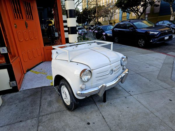 A white, vintage car front repurposed as a decorative piece is placed on the sidewalk near an orange building entrance.