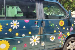 A blue van decorated with colorful flower stickers is parked on a sunny street near trees and a building.