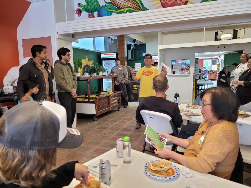 A group of people gather in a room with food, while one person in a yellow shirt stands and speaks, holding up a paper. Others are seated at tables, listening and eating.