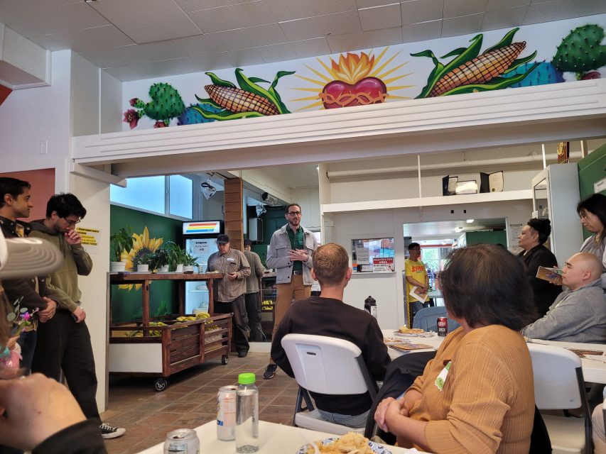 A group of people sit and stand in a community room as a man speaks at the front; colorful corn-themed art decorates the wall above him.